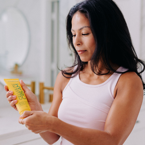 A woman with black hair pictured holding Frownies cellulite cream bottle in her hands and in the act of opening the top to use it 