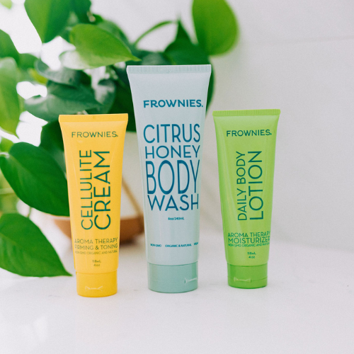 Frownies cellulite cream, citrus honey body wash, and daily body lotion pictured lined up next to each other in that order (left to right). There is a plant in the background