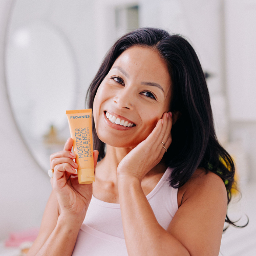 Frownies moisturizer for face and neck being held by a woman who is smiling, and is also rubbing the side of her face showing where you can apply the moisturizer 