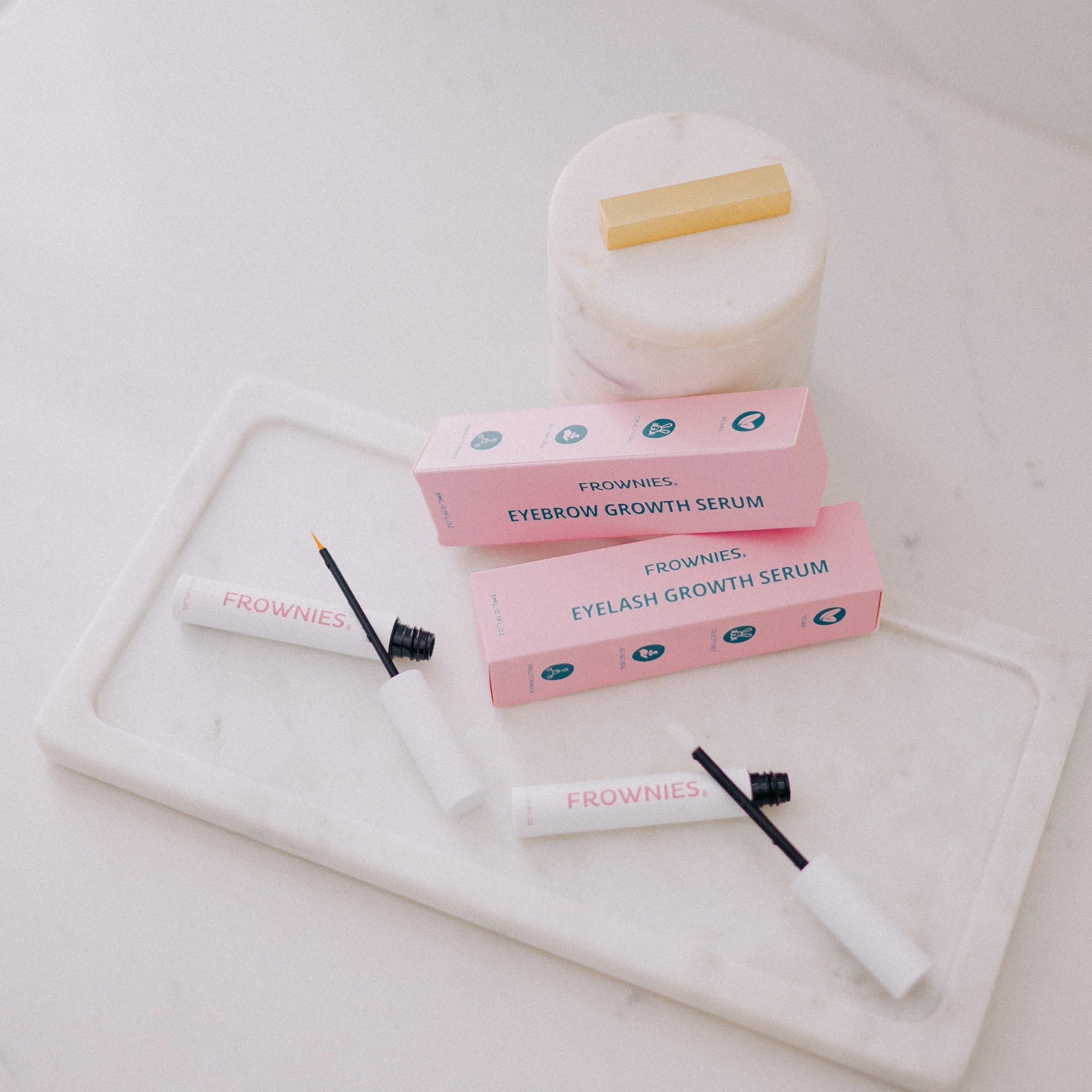 Frownies Eyelash Growth Serum and Eyebrow Growth Serum displayed together outside their packaging on a white tray, with the pink Frownies packaging also shown.