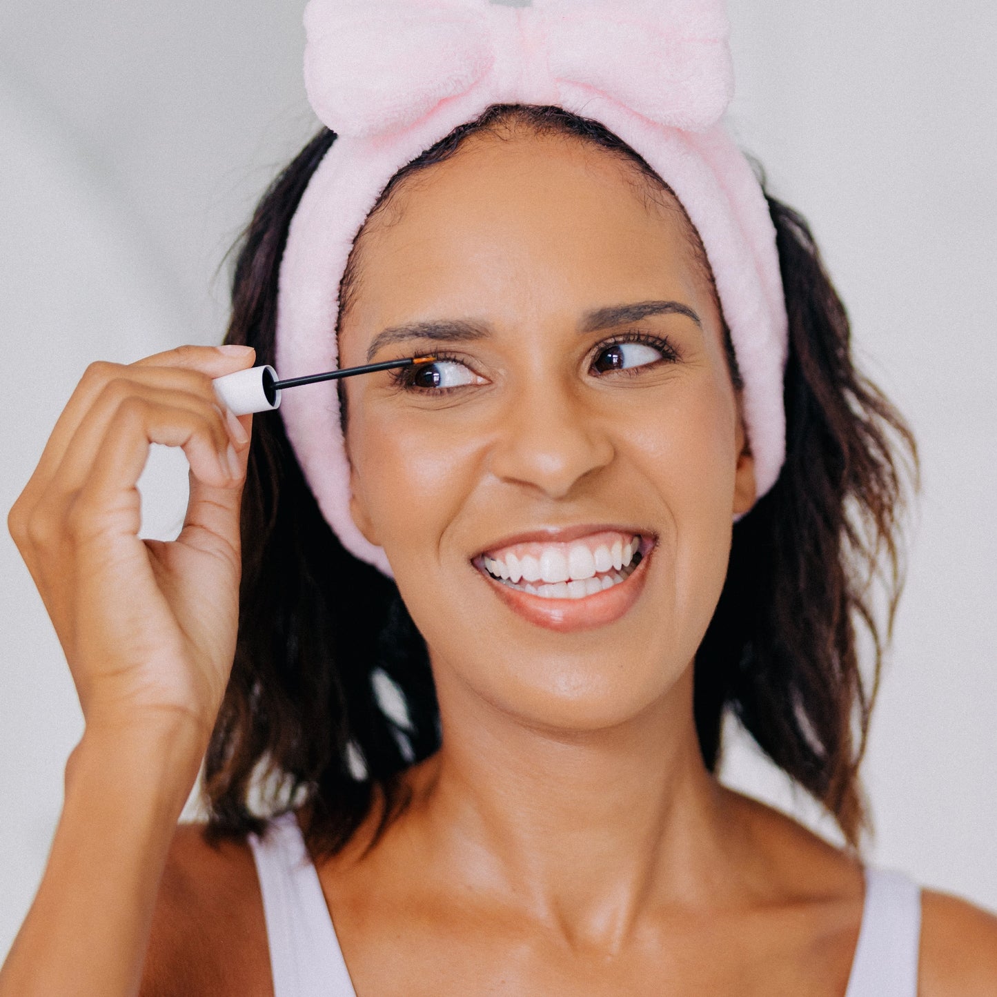 A happy woman applying Frownies Eyelash Growth Serum while wearing a Frownies skincare headband