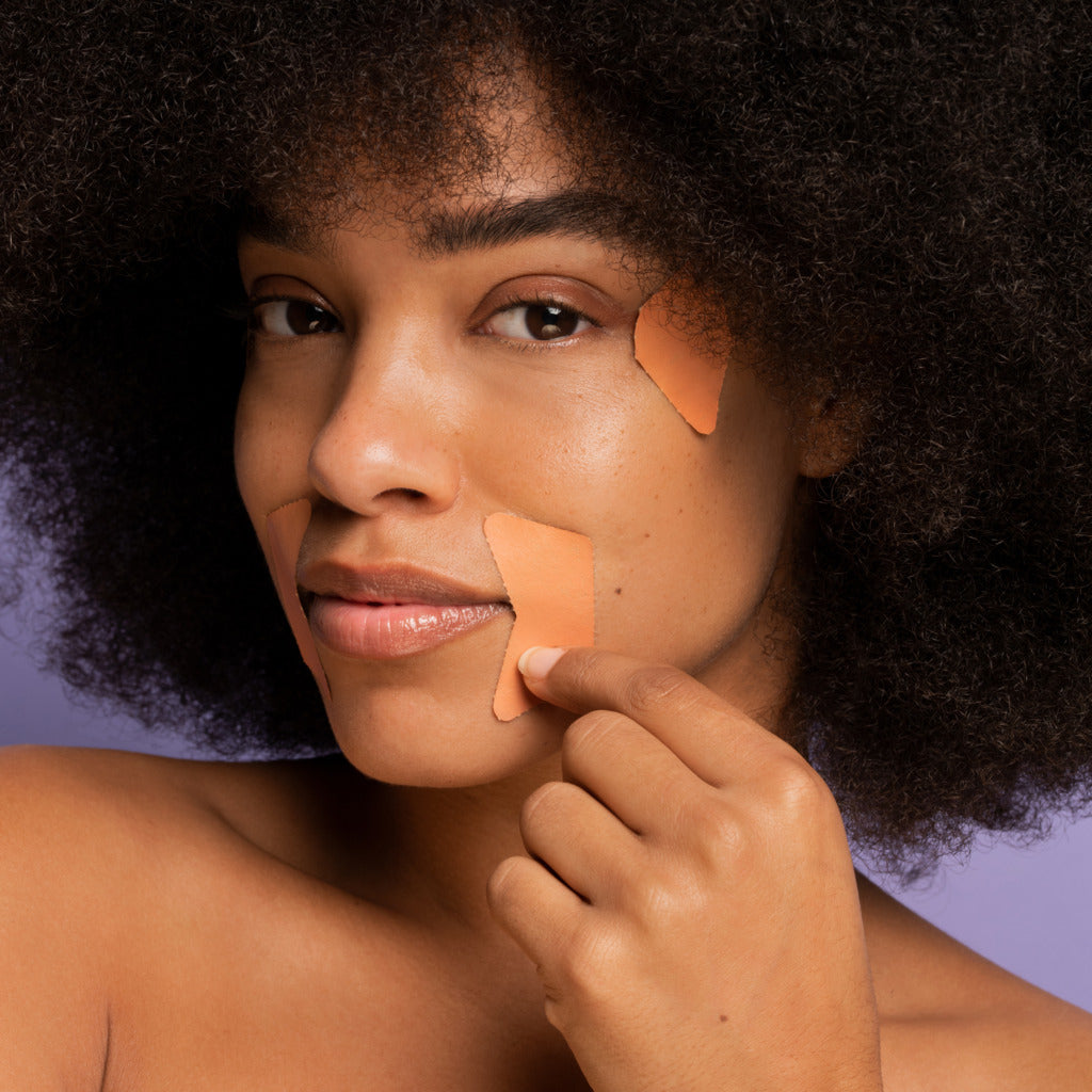 A black woman shown wearing Frownies corners of eyes and mouth facial patches to smooth expression lines. The picture has a lilac background