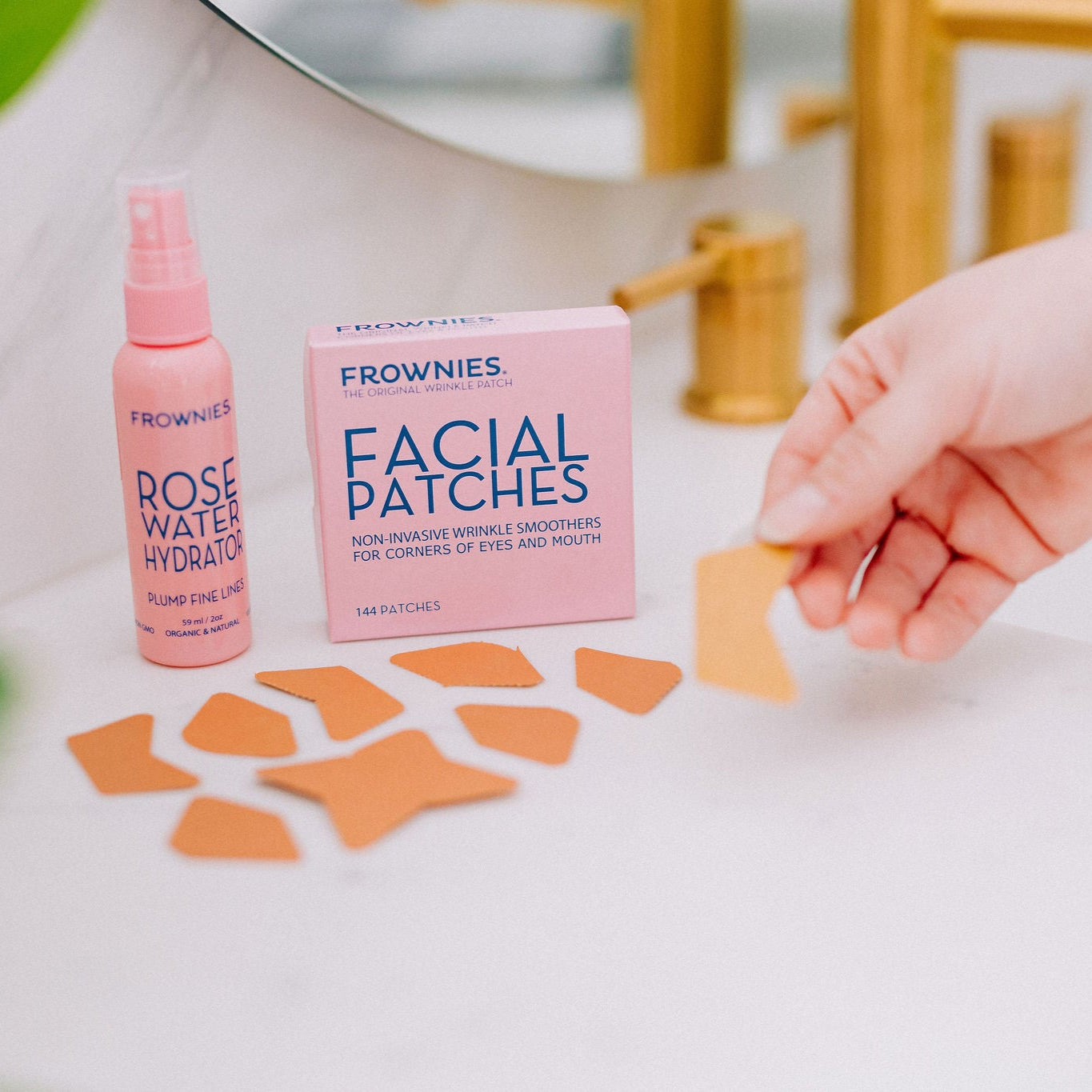 Frownies facial patches (CEM) and rose water hydrator spray shown sitting by a sink, while the facial patches are also laying on the sink counter top next to the packaging 