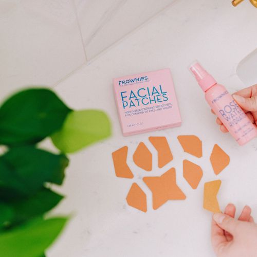 Frownies Corners of Eyes and Mouth (CEM) box displayed on a white surface with the patches spread out across the table. A hand is holding a bottle of Frownies Rose Water Hydrator spray in the picture showing they can be used together. 