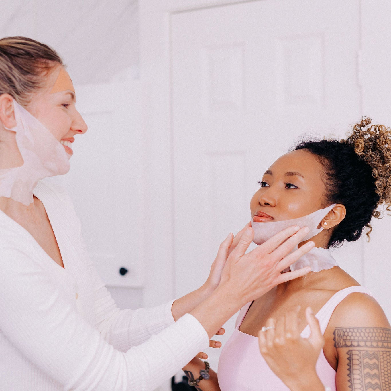 A black woman with curly hair having someone else put on her Frownies chin up polypeptide mask, the other woman is also wearing one