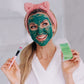 A woman wearing the frownies skincare headband shown with the frownies blue green algae clay mask on her face while holding the clay mask bag and brush up