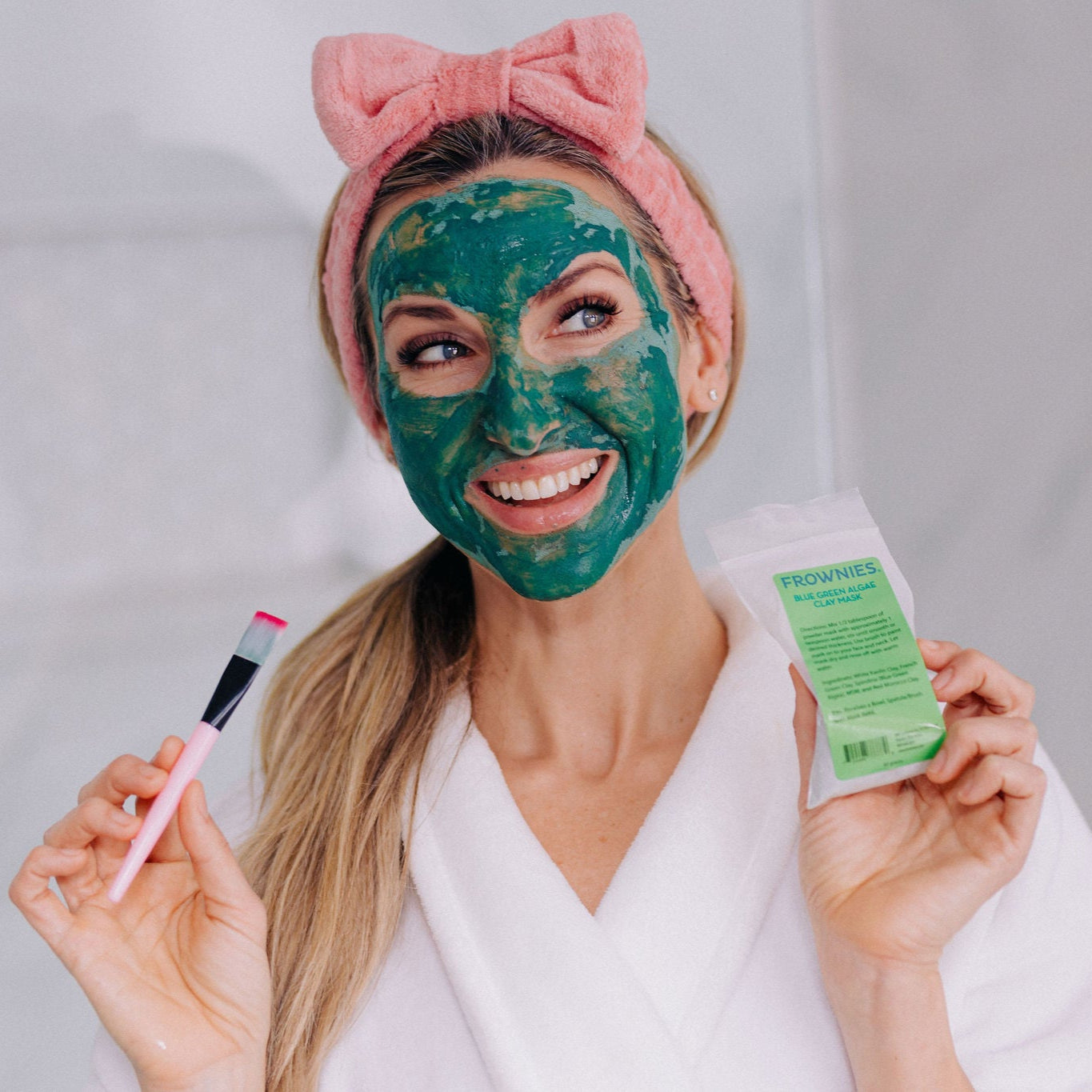 A woman wearing the frownies skincare headband shown with the frownies blue green algae clay mask on her face while holding the clay mask bag and brush up