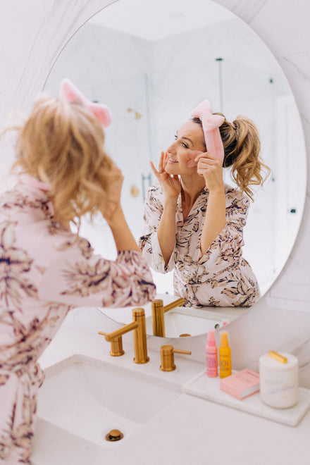 A woman shown in the mirror wearing frownies skincare headband and using frownies gua sha rose quartz stone on the cheek area of her face