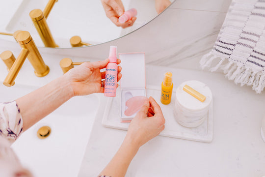 Frownies rose water hydrator spray shown being held in one hand while the gua sha rose quartz stone in the other hand. This picture is by a bathroom sink