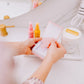 Frownies gua sha rose quartz stone being taken out of the box by a woman, the picture only shows her hands. Frownies Rose water hydrator spray and skin serum are also seen in the background of the picture 