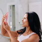 A woman with black hair shown holding Frownies rose water hydrator spray up while she looks at the bottle. It looks like she is about to use it and spray it onto her face. 