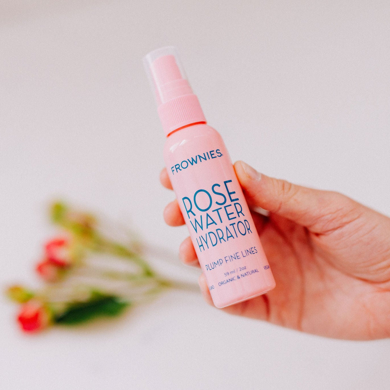 Frownies rose water hydrator spray that plumps fine lines shown being held with actual roses blurred out in the background