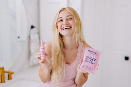 A woman with blonde hair smiling and shown holding Frownies under eye gels in one hand and rose water hydrator spray in the other hand