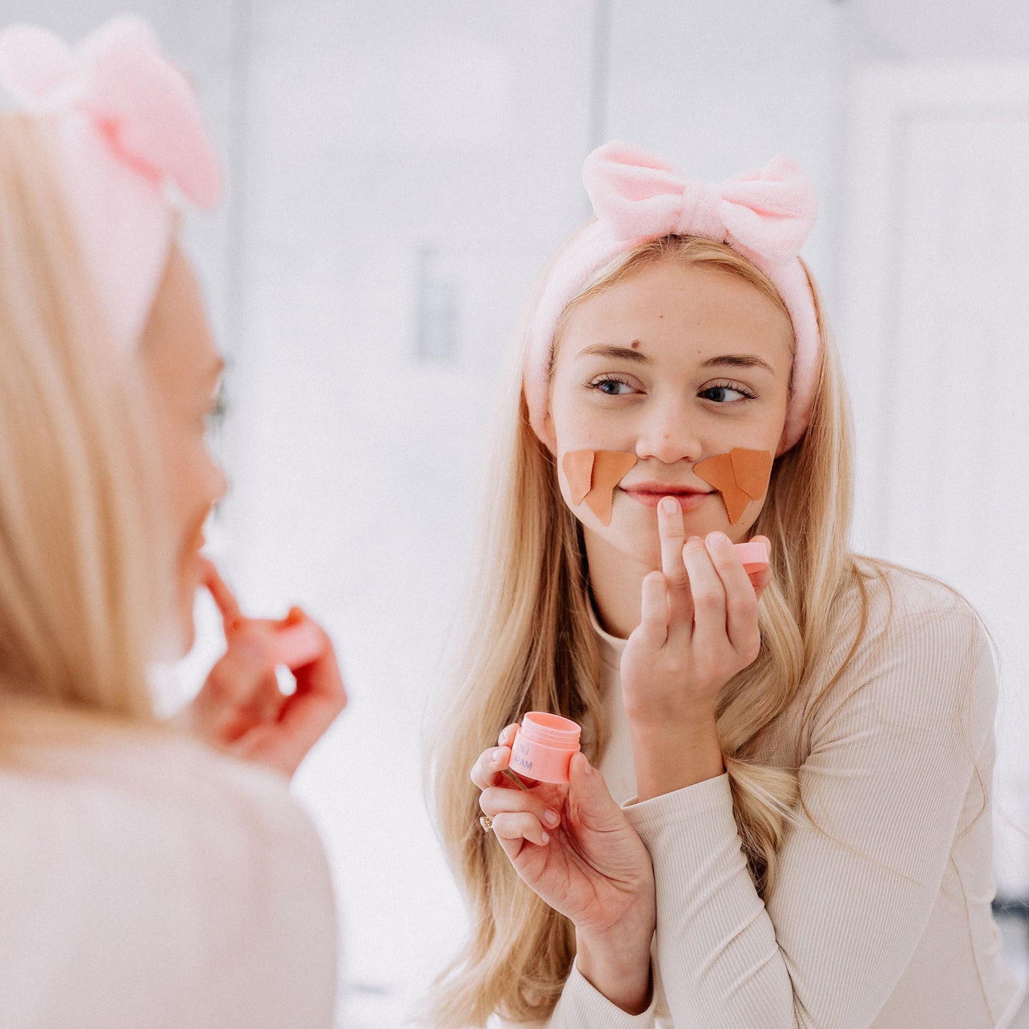 A woman with blonde hair wearing the Frownies skincare headband while applying the Frownies lip mask to her lips 