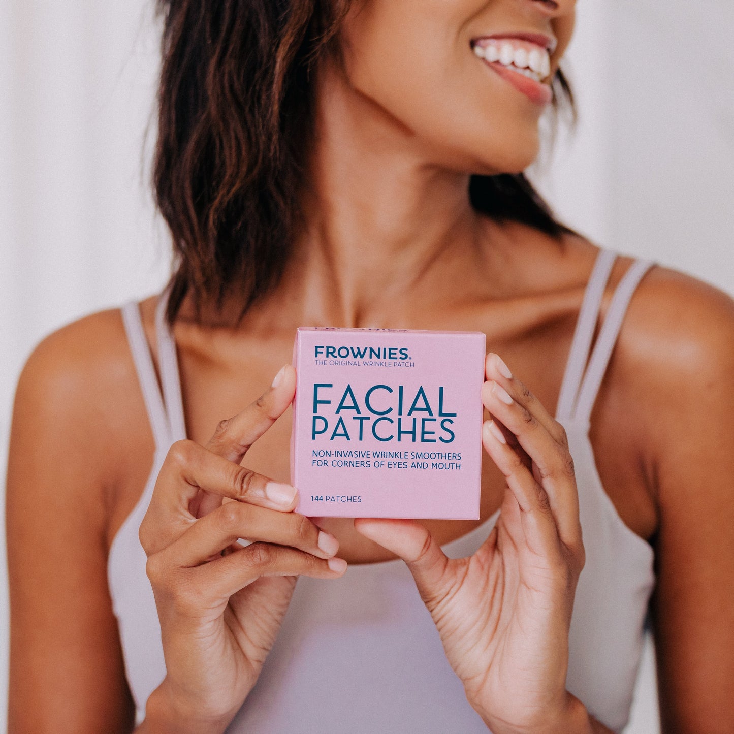 A black woman with black hair shown holding Frownies Corners of eyes and mouth facial patches while smiling 