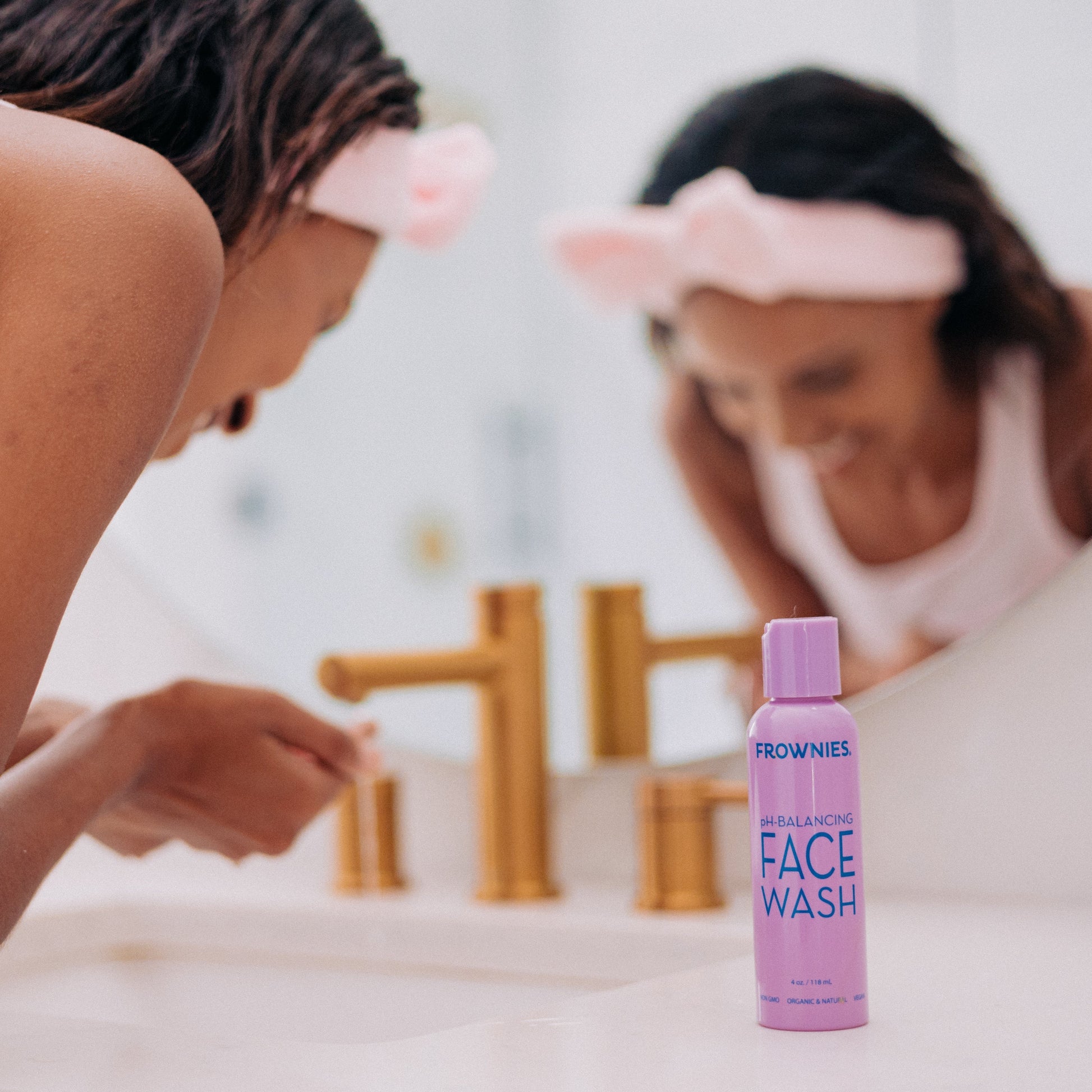 A woman in the bathroom sink washing her face while Frownies pH-balancing face wash is beside her on the counter top, she is also wearing Frownies skincare headband