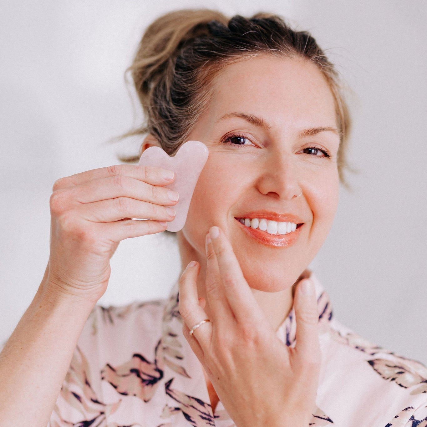 Frownies Gua Sha rose quartz stone being used by a white woman with dirty blonde hair. She is using it on her right check area