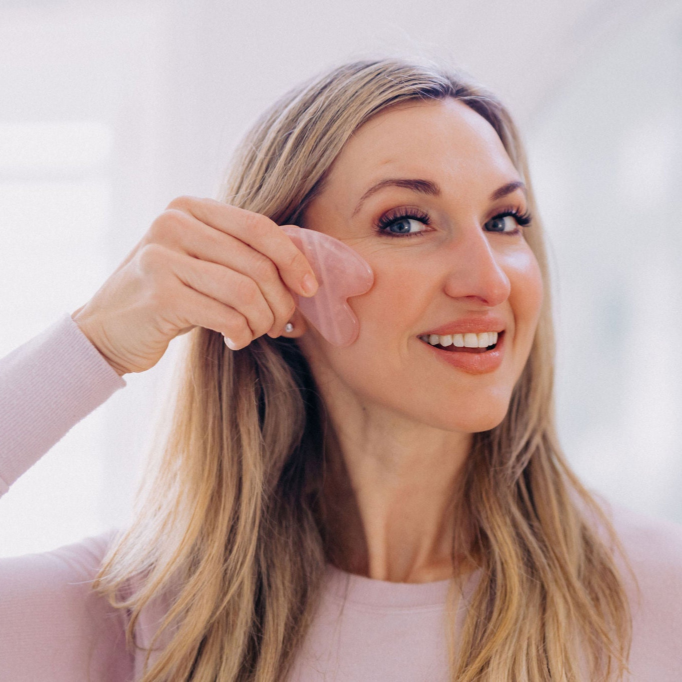 Frownies Gua Sha rose quartz stone being used by a white woman with dirty blonde hair. She is using it on her right check area