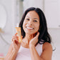 A woman pictured smiling and holding Frownies moisturizer for face and neck while also applying some to her neck