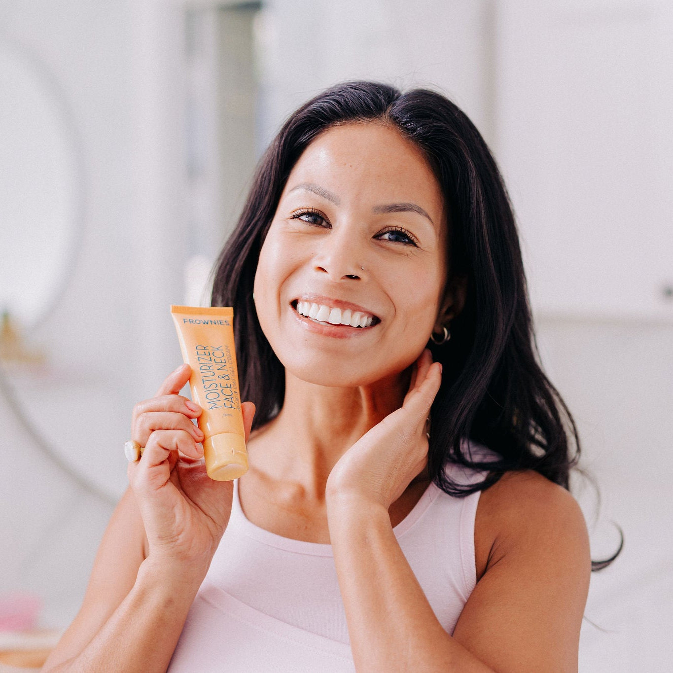 A woman pictured smiling and holding Frownies moisturizer for face and neck while also applying some to her neck