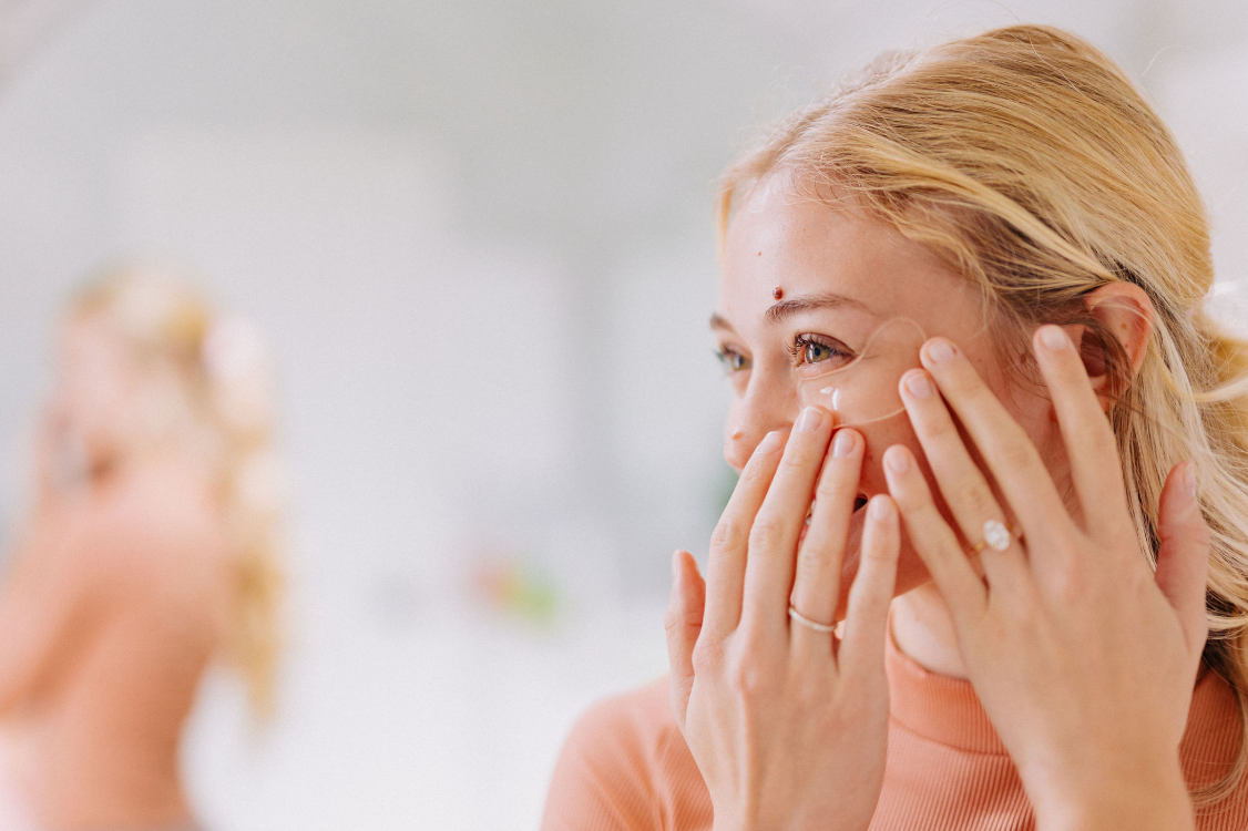 young blonde woman applying frownies under eye gels in front of a mirrorr