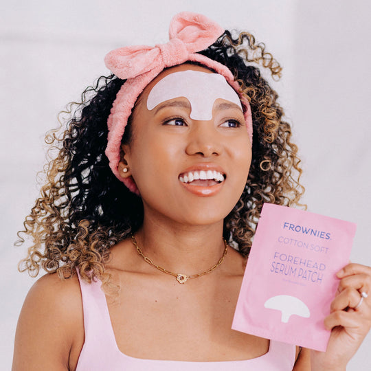 A happy woman with curly hair, smiling while wearing a Frownies forehead serum patch and a Frownies skincare headband, holding up the forehead serum patch packaging for the camera to see.