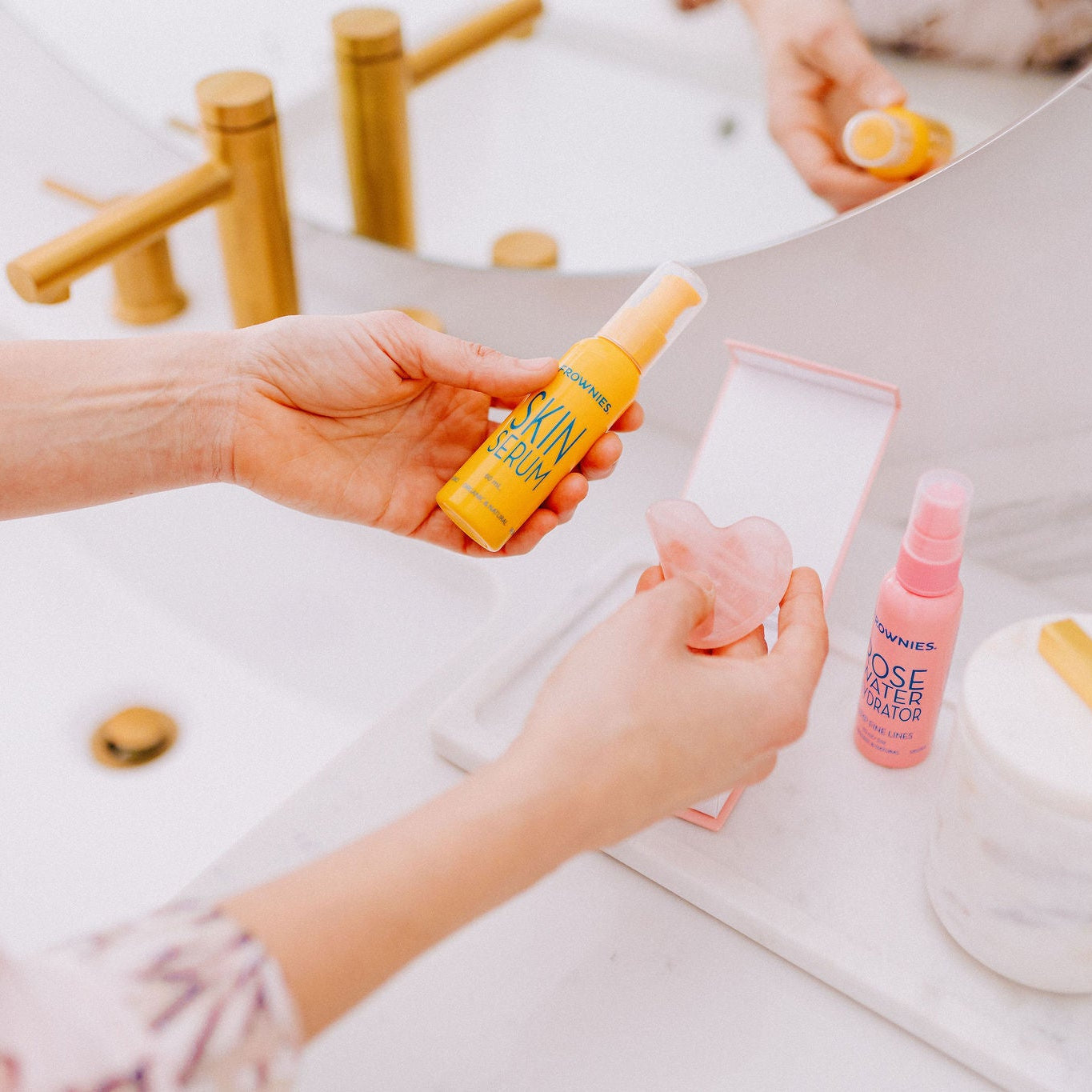 Frownies skin serum bing held over a bathroom sink with the gua sha rose quartz stone also being held in the other hand. Frownies rose water hydrator spray is also shown in the photo as well 