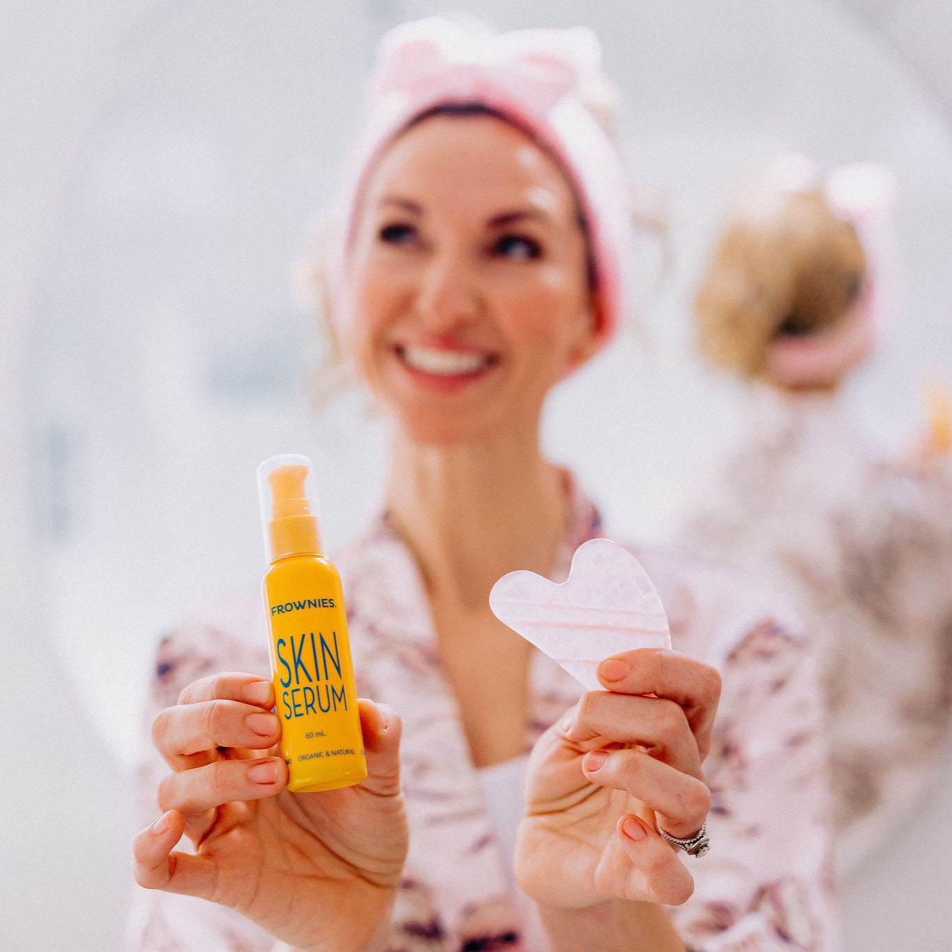 Frownies skin serum and gua sha rose quartz stone being held by a woman wearing the Frownies skincare headband. The woman is smiling and looks cheerful