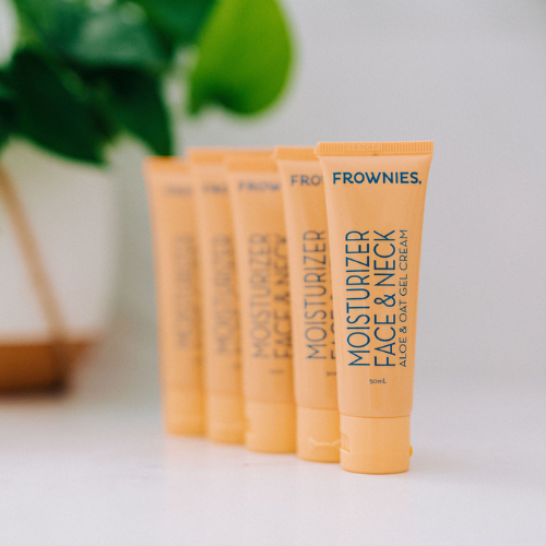 5 Frownies face and neck moisturizer with aloe and oat gel lined up diagonally, with some blurred out plants in the background