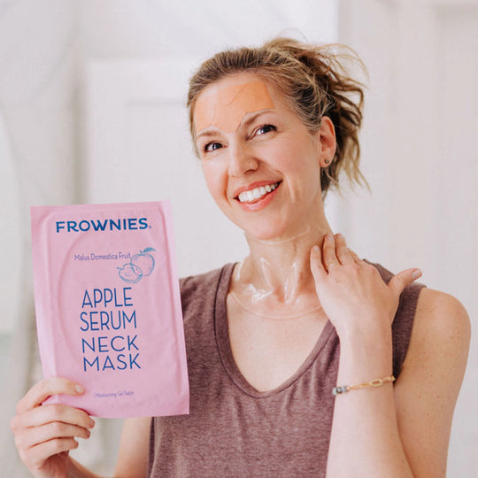 A woman smiling while wearing an apple neck mask, holding up the Frownies packaging for the camera to see.