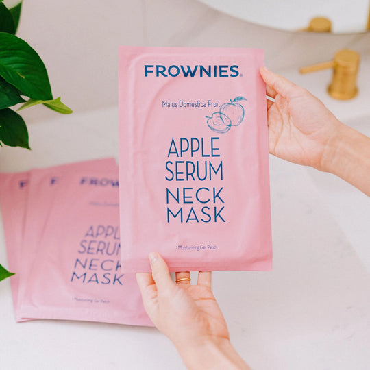 A woman’s hands holding a Frownies apple serum neck mask, with additional masks laying on the table below.