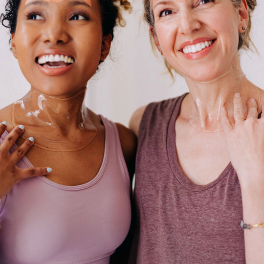 Two women smiling with their shoulders touching, both wearing apple neck masks and demonstrating how to wear them