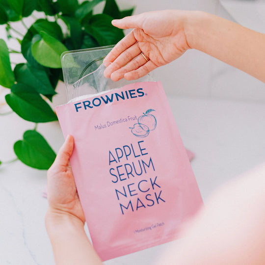 A pair of hands taking a Frownies apple serum neck mask out of the packaging.