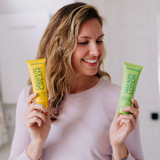 Woman with dirty blonde hair smiling and looking down and away from the camera, holding Frownies Daily Body Lotion and Cellulite Cream.