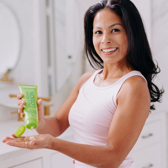 Woman with dark black hair smiling, holding Frownies Daily Body Lotion in her right hand while squeezing some into her left hand