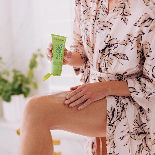 Close-up of a woman's leg in a robe while applying Frownies Daily Body Lotion.