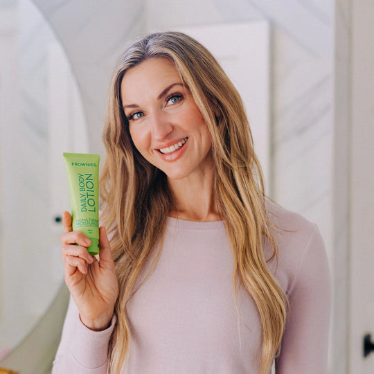 Blonde woman smiling with her head tilted, holding Frownies Daily Body Lotion.