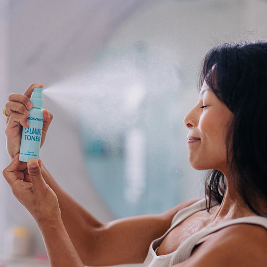 A woman spraying her face with Frownies calming toner, with the bottle and mist visibly spraying.