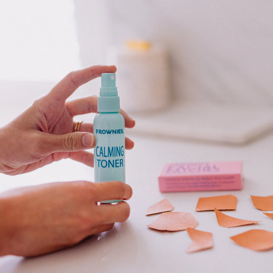 Hands holding the Frownies calming toner, with facial patches laid out around the bottle.