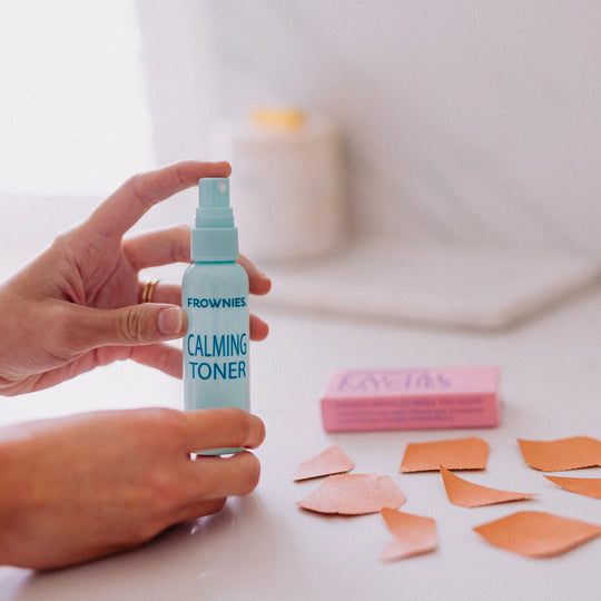 Hands holding a bottle of Frownies Calming Toner with facial patches laid out beside the bottle on a flat surface