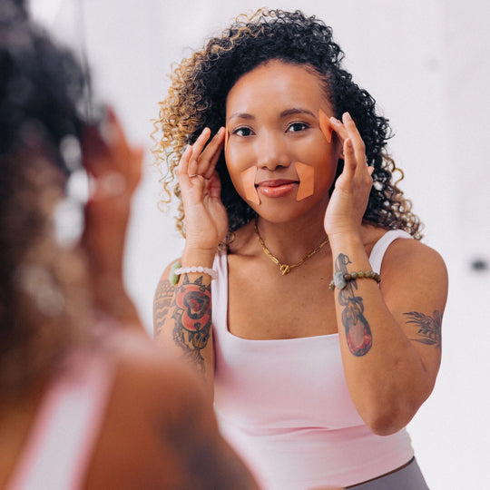 A woman demonstrating how to wear Frownies facial patches while looking in the mirror. She is wearing frownies CEM (corners of eyes and mouth).