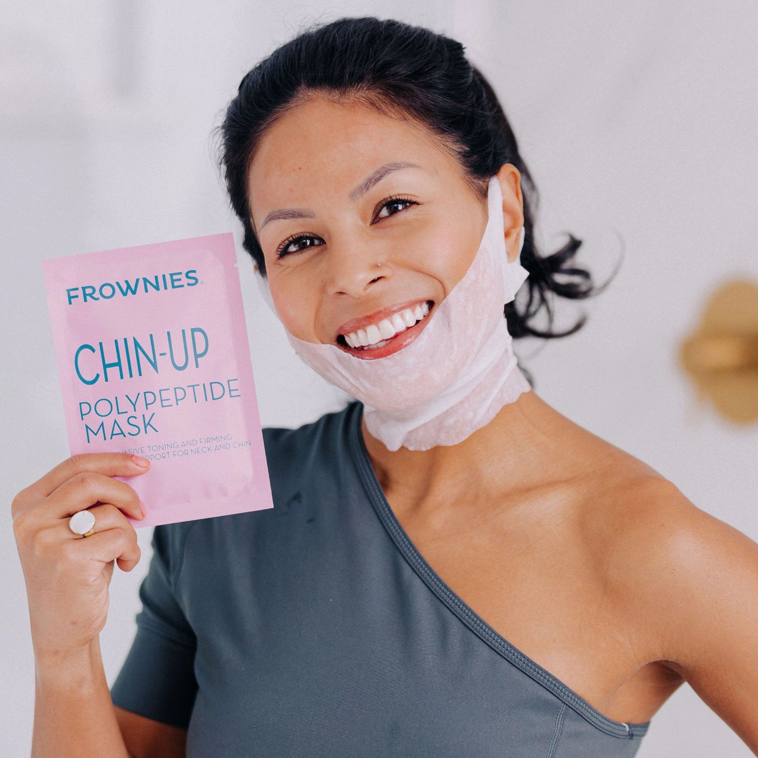 A woman with black hair smiling with her head tilted, wearing a Frownies Chin-Up Polypeptide Mask, and holding the packaging up to the camera.