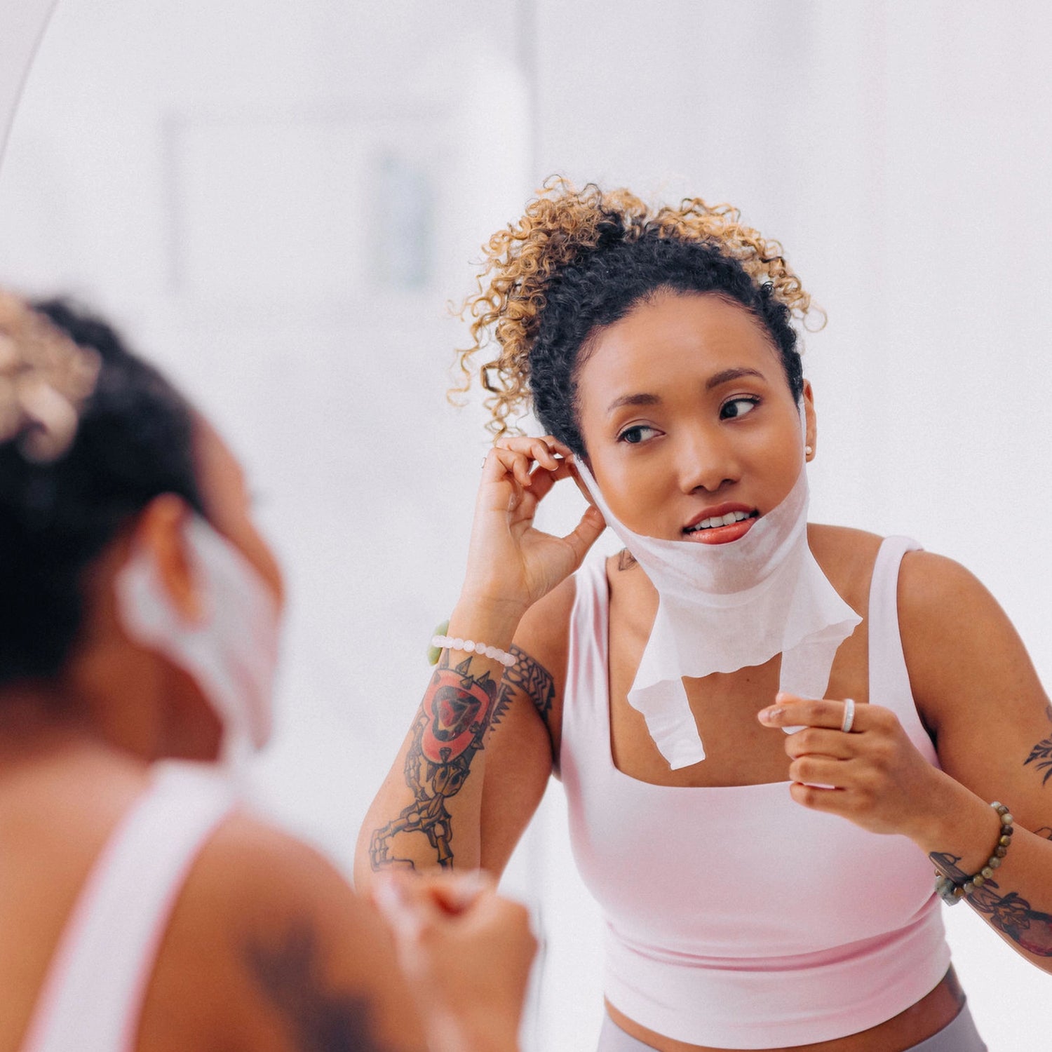 A woman with curly hair demonstrating how to put on the Frownies Chin-Up Polypeptide Mask in front of a bathroom mirror.