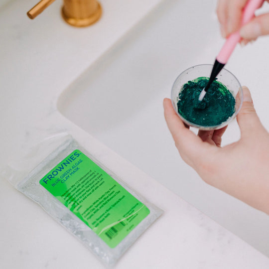 Frownies Blue Green Algae Mask shown out of its package, being mixed and prepared for use on a bathroom counter.