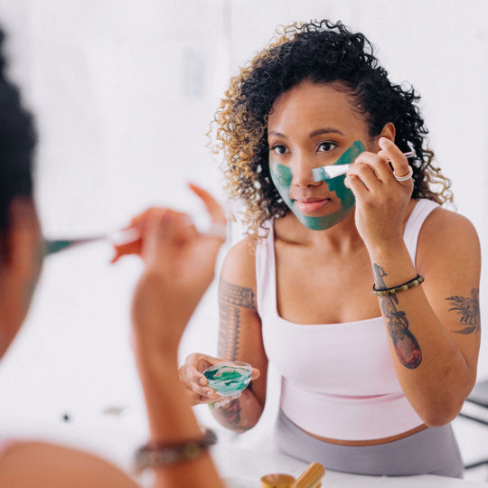 Curly-haired woman applying Frownies Blue Green Algae Mask in front of a bathroom mirror, holding a bowl in one hand and using a brush to apply the mask to her face.