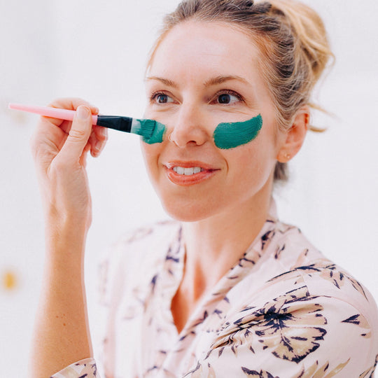 Close-up of a blonde woman in a bathroom mirror using a brush to apply her Blue Green Algae Mask.