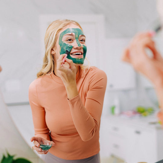 Blonde woman in an orange shirt applying Frownies blue-green algae clay mask to her face while looking in the mirror.