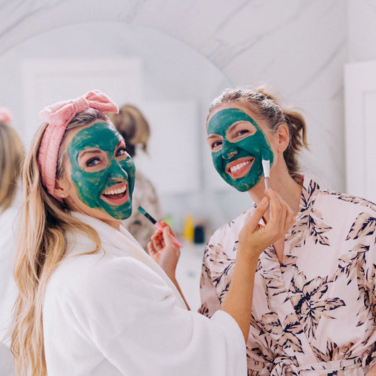 Two women joyfully applying blue algae clay mask to each other’s faces in a bathroom, with a mirror in the background. One is also wearing Frownies skin care headband. 