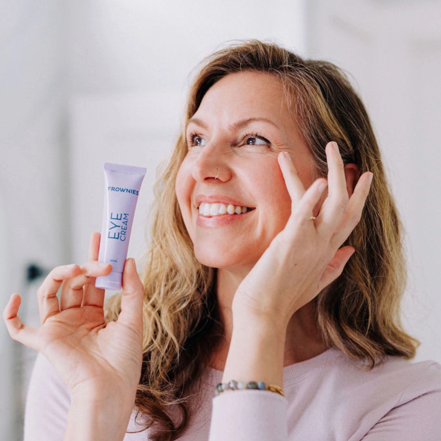 Woman smiling and slightly looking away while carefully touching her eye, holding Frownies Eye Cream in her hand and showcasing the product.