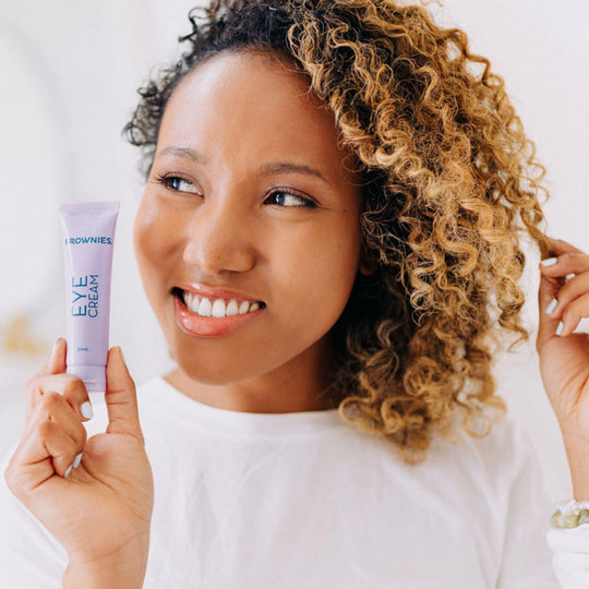 Woman with curly hair smirking and looking away from the camera, twirling her hair while holding Frownies Eye Cream in the other hand, clearly showcasing the product.